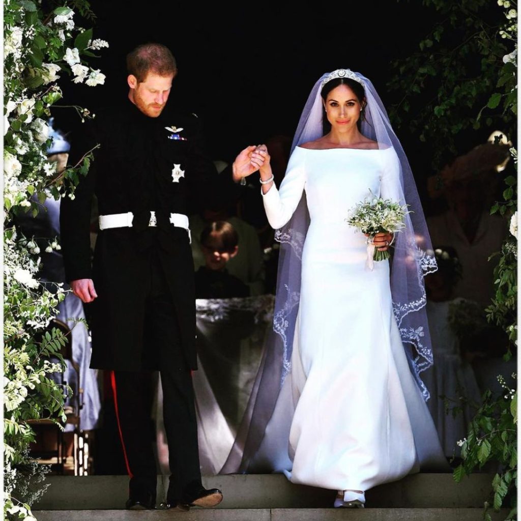 Na imagem há Meghan Markle vestida de noiva, Príncipe Harry segura sua mão enquanto a duquesa de Sussex sorri descendo uma escadaria