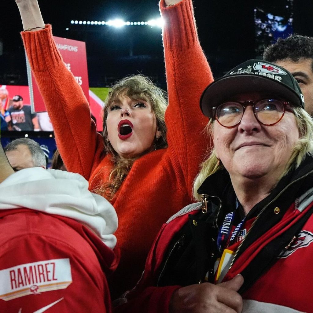 Taylor Swift aparece com camisa vermelha, braços estendidos em uma típica comemoração