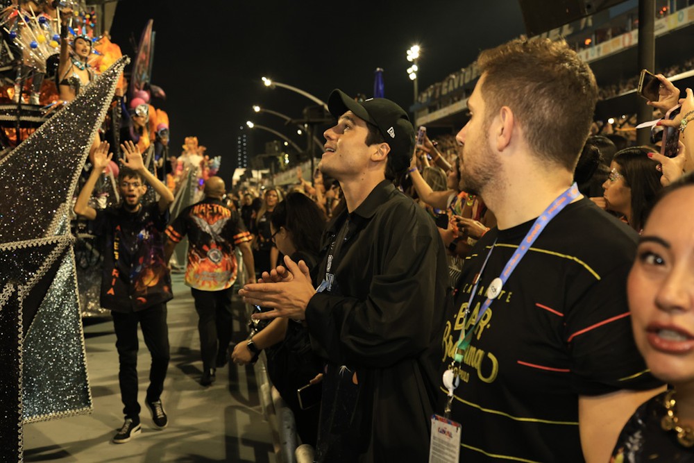 Na imagem aparece Nicolas Prattes admirando o Desfile das Campeãs. Ao lado há Karina Sato, irmã da apresentadora