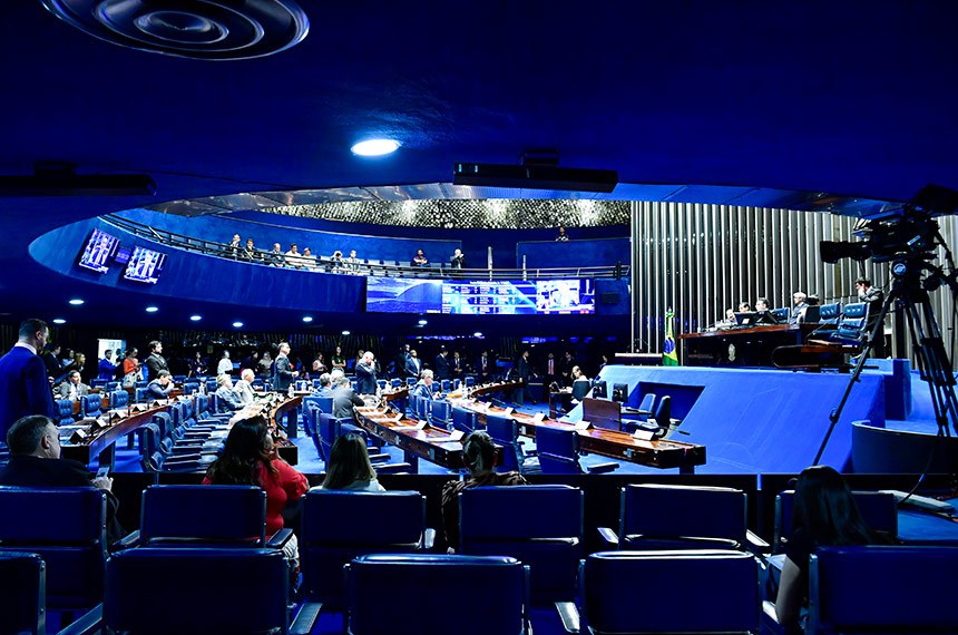 Na imagem diversas cadeiras azul em formato circular compõe a sala do senado brasileiro. Acima há um mezanino que segue o mesmo critério de organização. Nas cadeiras os senadores se reúnem para discutir projeto de lei.