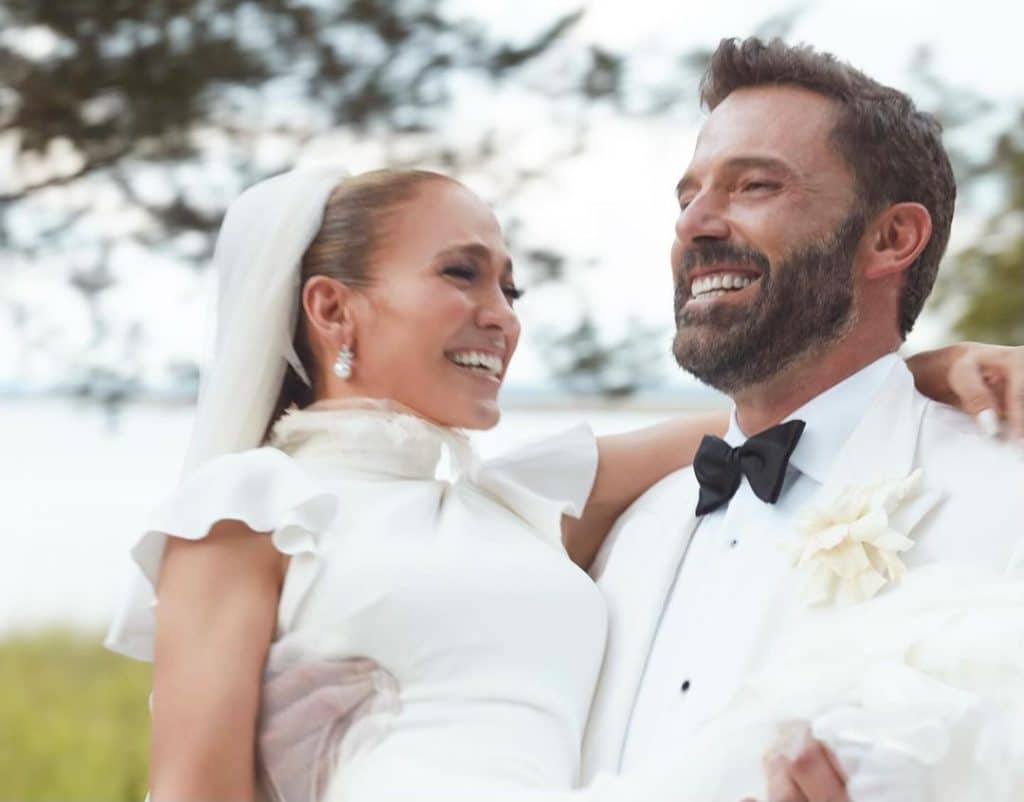 Jennifer Lopez e Ben Affleck sorridentes em foto durante casamento. Jennifer está com um vestido branco que vai até o pescoço e tem babados nas mangas, com o véu para trás. Ben veste um terno branco com uma gravata borboleta preta. O casal está em um jardim.