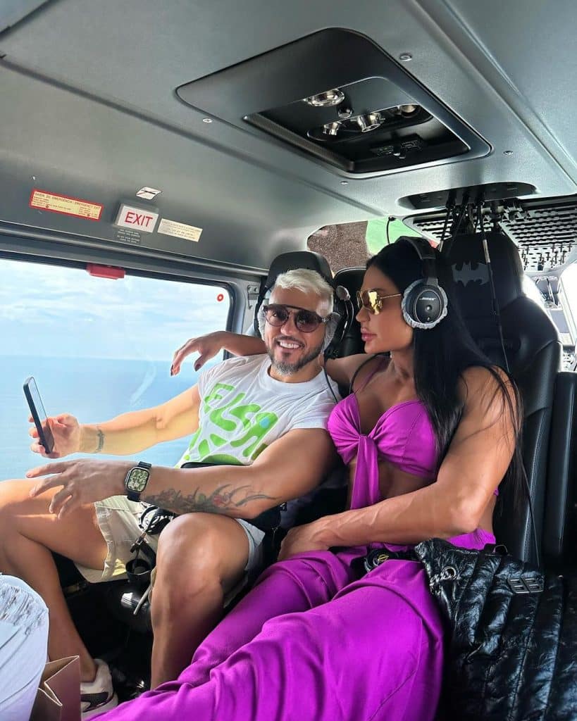 Belo e Gracyanne Barbosa em passeio de helicóptero. Belo está na esquerda, vestindo bermudas e blusa branca, já Gracyanne está vestindo conjunto roxo.