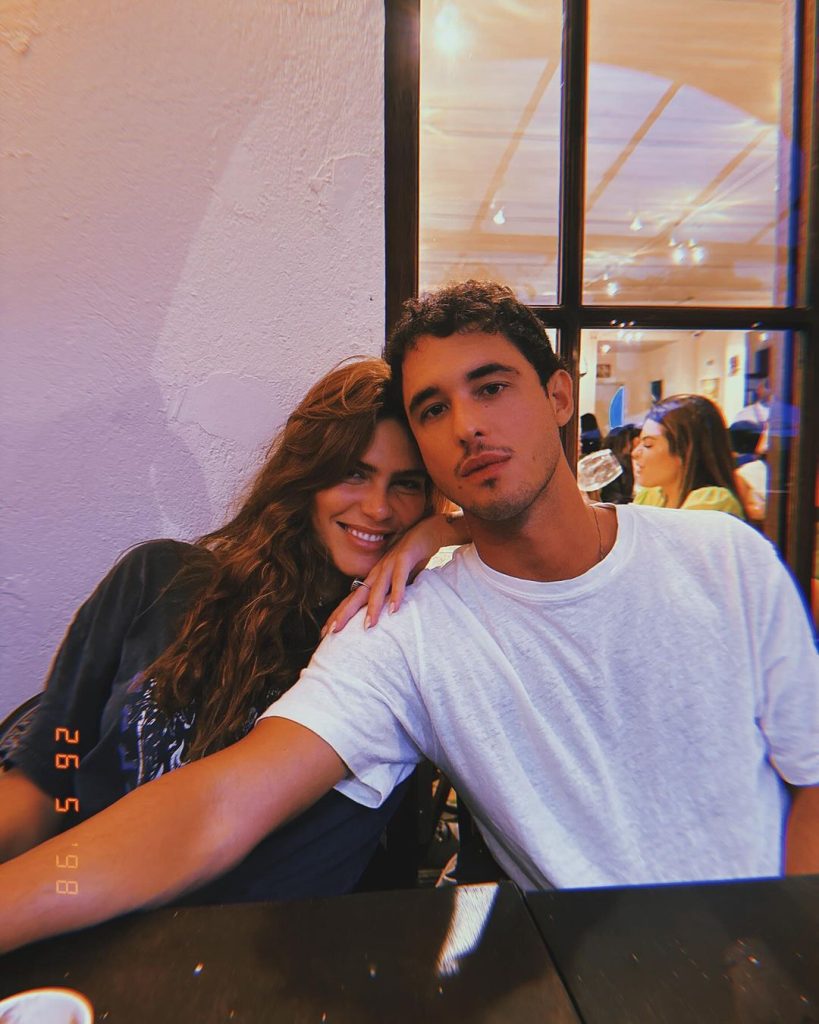 Mariana Goldfarb e Rafael Kemp posam atrás de mesa de restaurante. Ela está apoiada em seu braço e veste uma jaqueta de couro preta, com os cabelos soltos. Rafael veste uma camiseta branca.