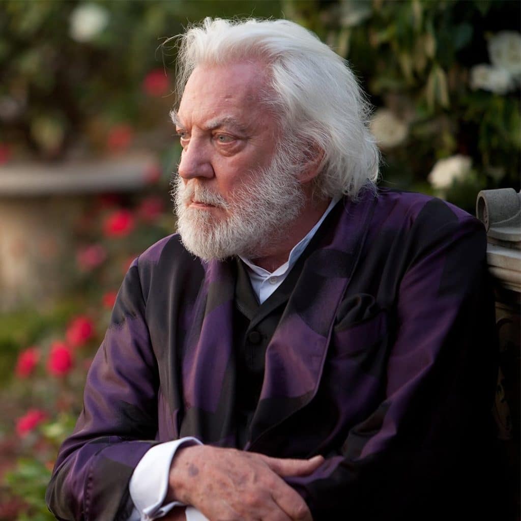 Donald Sutherland caracterizado como presidente Snow, de barba e cabelos brancos; usa roupa roxa e está sentado olhando para algo fora do ponto de vista da câmera.